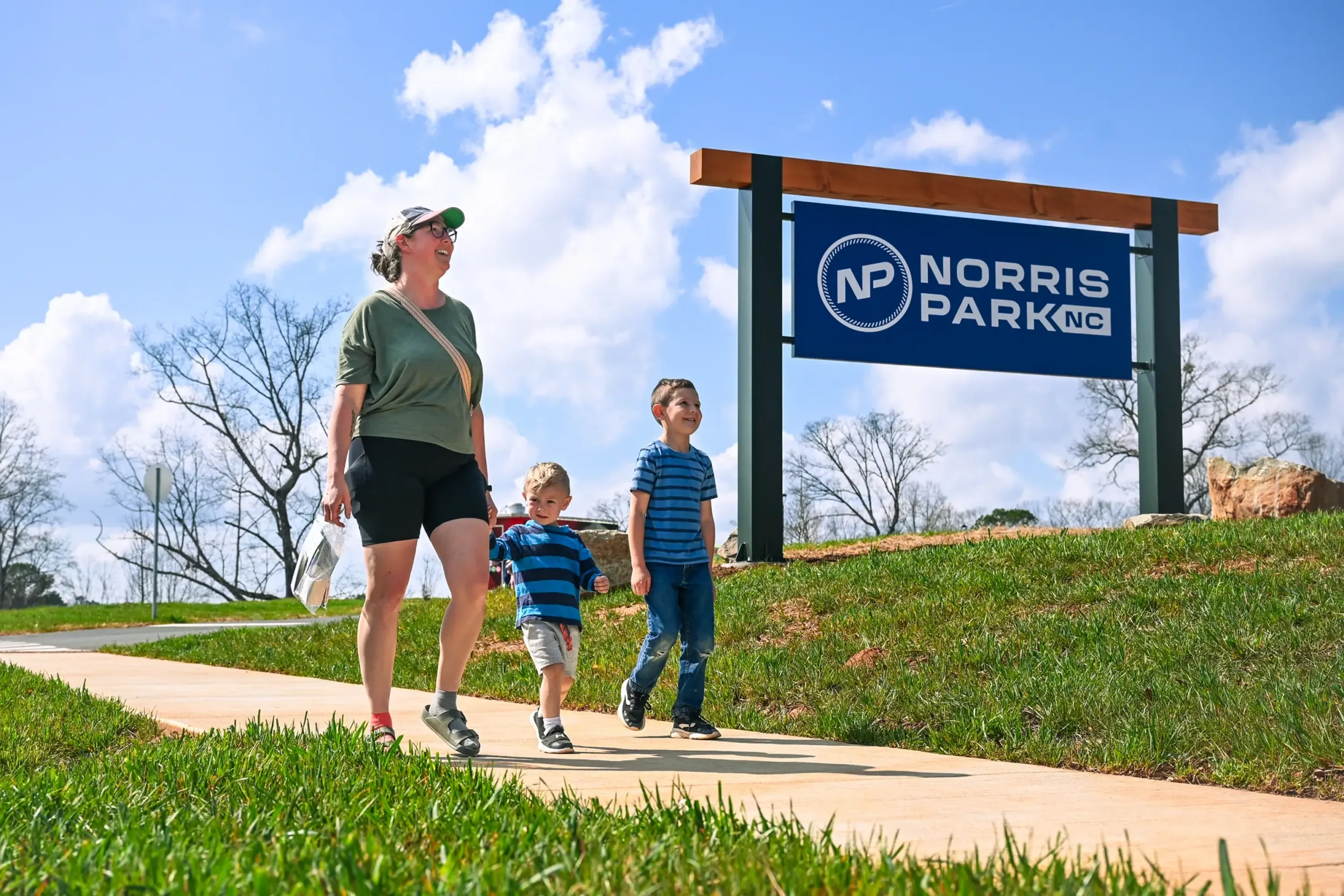 A mother with two kids walking down the sidewalk by the Norris Park entrance sign
