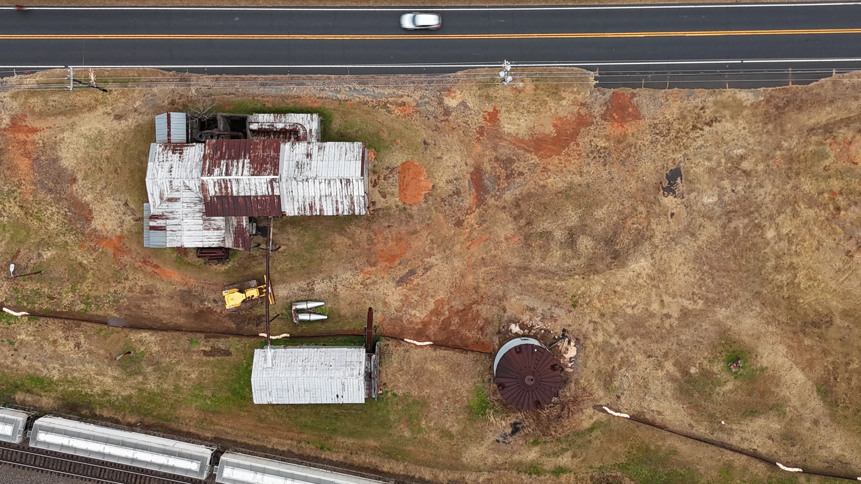 Aerial view of the Colfax Cotton Gin site with rusted sheds, a round tank and a rail line beside a highway.